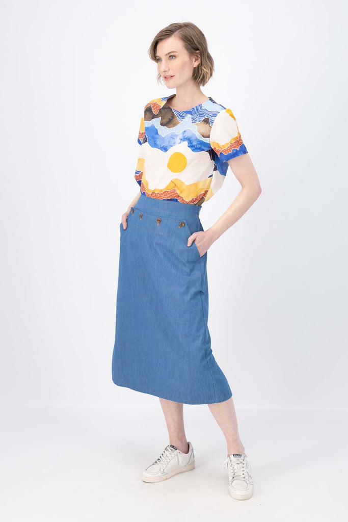 Olga de Polga Hepburn skirt in light wash cotton denim. The Hepburn Skirt is a chic pencil skirt with a playful twist. Featuring a unique button detail at the waist that forms a flattering pocket shape. Side view on model.