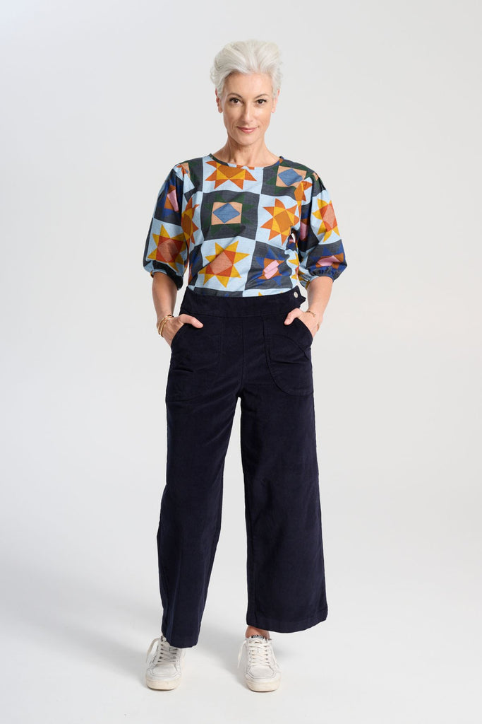 The blouse features a beautifully detailed lantern sleeve with a drop shoulder and delicate pleating on the shoulder and cuff. It also has a wide boat neckline and invisible zip fastening at the back. Front full view with navy pants.