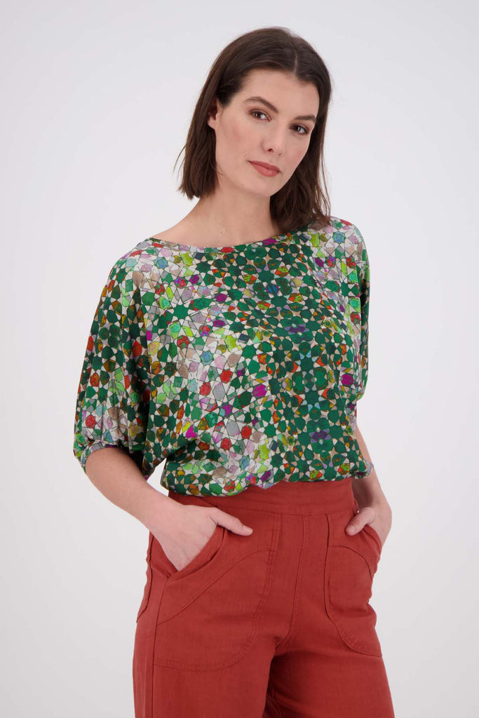 Olga de Polga Kaleidoscope Green Camisa Blouse in printed fine Tencel. This is a reversible top with the choice of a soft V-neck and a boat neck which are interchangeable. Half length sleeves. Finishes at the hip. Side front close up view on model.