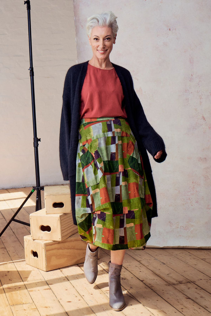 Olga de Polga Sage green Kismet printed Milwaukee skirt in fine cord. Large patch pockets. Unique panelled design. Back elasticated waistband. Front view, worn with a navy knit cardigan.