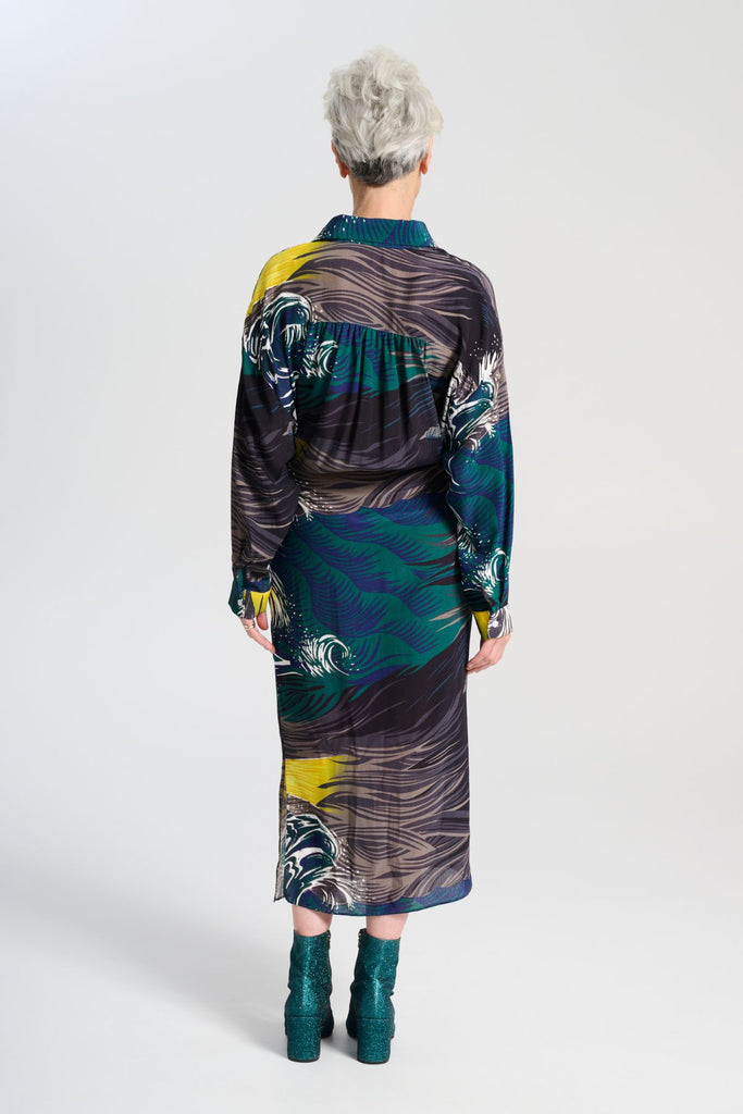 Olga de Polga Fluido wrap shirt dress. Printed Viscose. The Isla print captures the power of nature, with crashing waves and a guiding lighthouse. Deep blues, stormy greens, and yellow accents evoke hope and direction through turbulent times, reflecting the challenges of life and the human drive to navigate the unknown. Back view on model