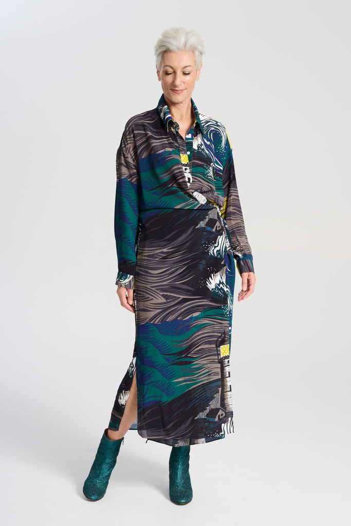 Olga de Polga Fluido wrap shirt dress. Printed Viscose. The Isla print captures the power of nature, with crashing waves and a guiding lighthouse. Deep blues, stormy greens, and yellow accents evoke hope and direction through turbulent times, reflecting the challenges of life and the human drive to navigate the unknown. Front full length view on model