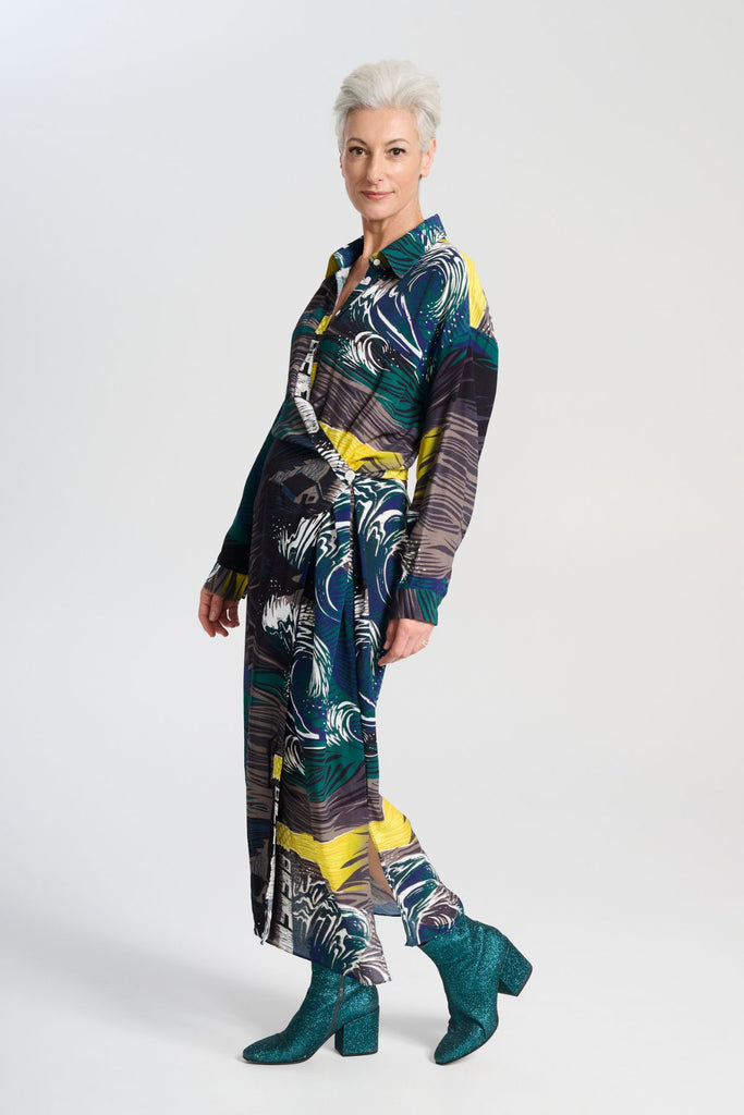 Olga de Polga Fluido wrap shirt dress. Printed Viscose. The Isla print captures the power of nature, with crashing waves and a guiding lighthouse. Deep blues, stormy greens, and yellow accents evoke hope and direction through turbulent times, reflecting the challenges of life and the human drive to navigate the unknown. Side view on model