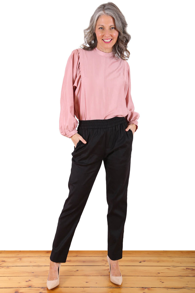The Hamptons trousers in black. The perfect mixture of comfort and class, the Hamptons Trousers will be your next throw-on-and-go pants for any situation - from the office to the beach. These pants are high waisted, with a straight fit around the waist and thighs that then tapers to the ankle. The elasticated waistband also includes a hidden drawstring so you can loosen and tighten the pants as you desire. Front view.